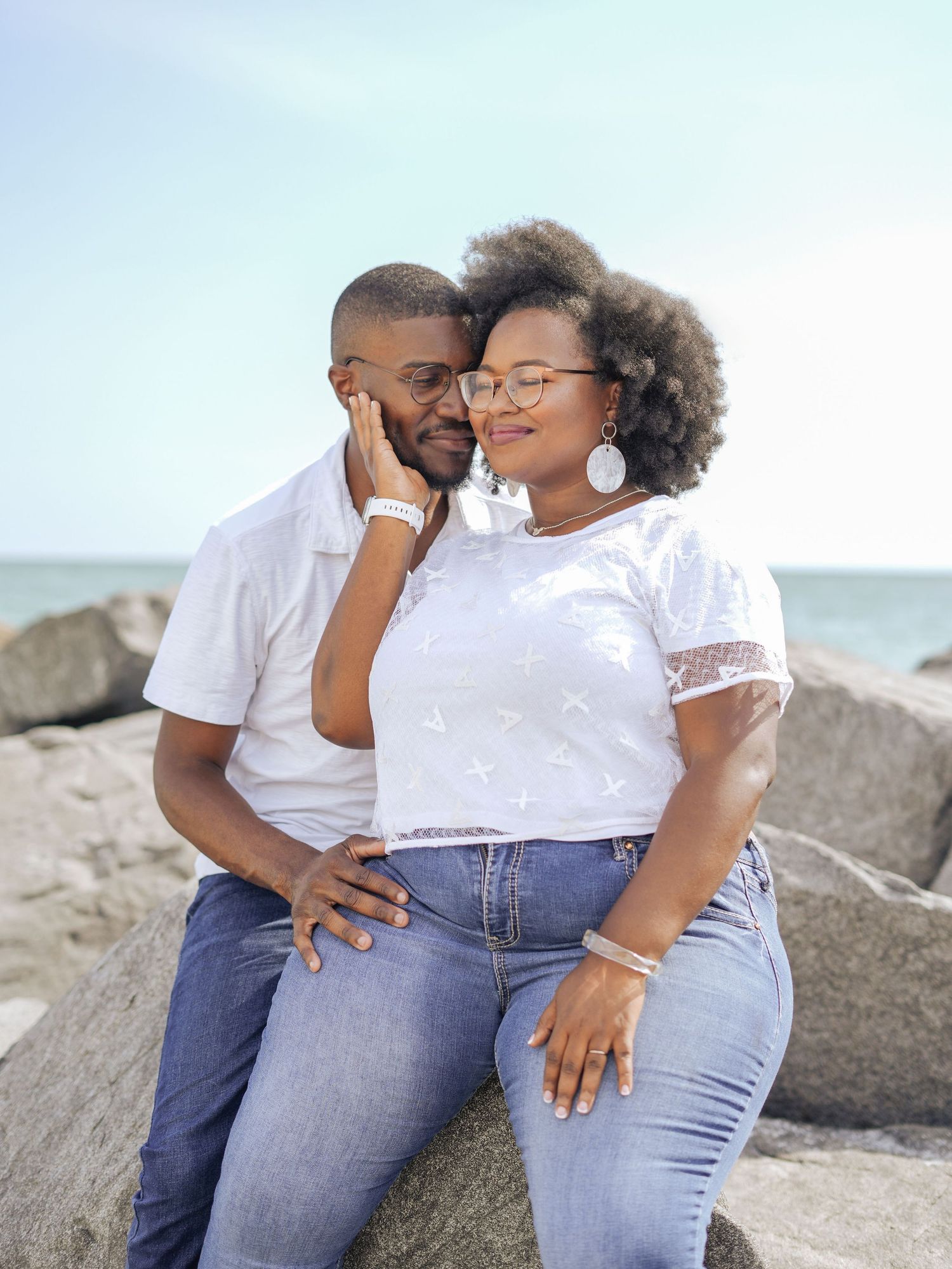 A couple in matching white tops and jeans share a tender moment sitting on rocks by the ocean.