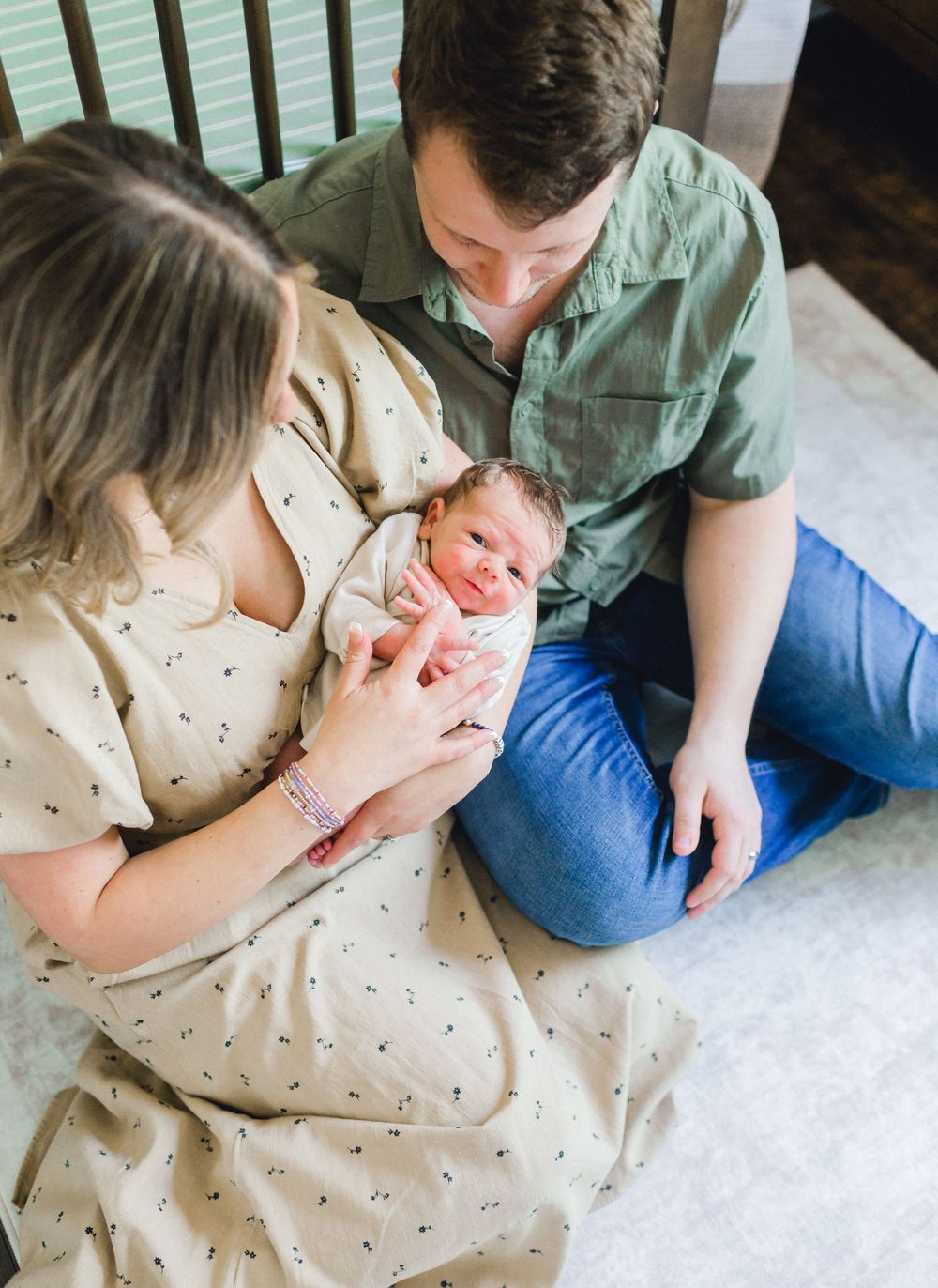 New parents sitting on floor gazing lovingly at their swaddled newborn baby during an in-home photo session.