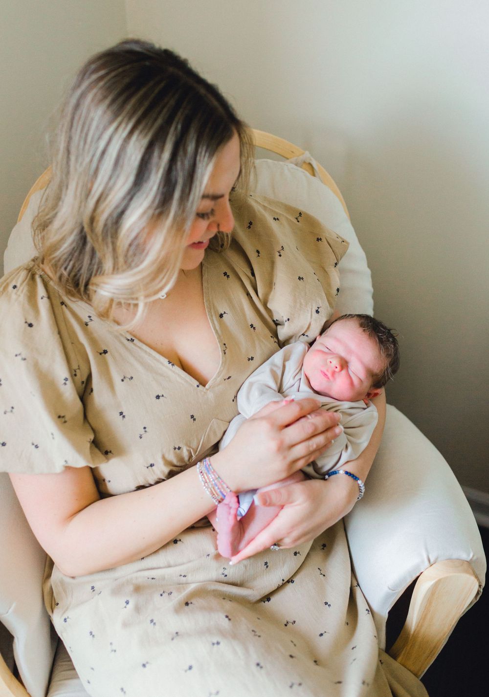 A new mother in a tan floral dress cradles her sleeping newborn baby while seated in a wooden rocking chair.