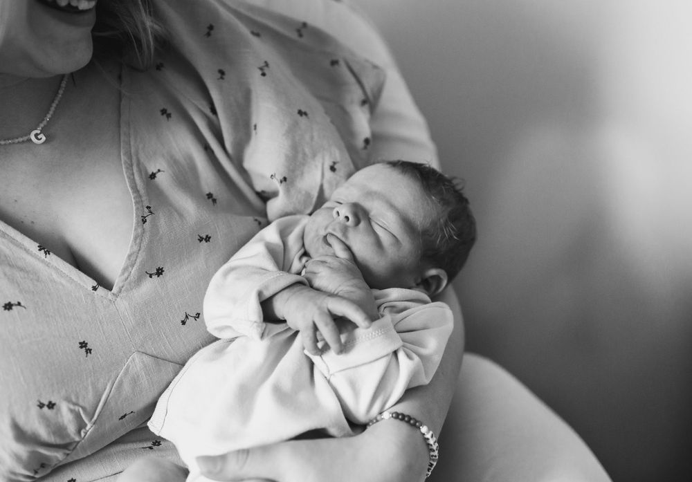 Black and white photo of a newborn baby sleeping peacefully in mother's arms, wearing a white onesie.