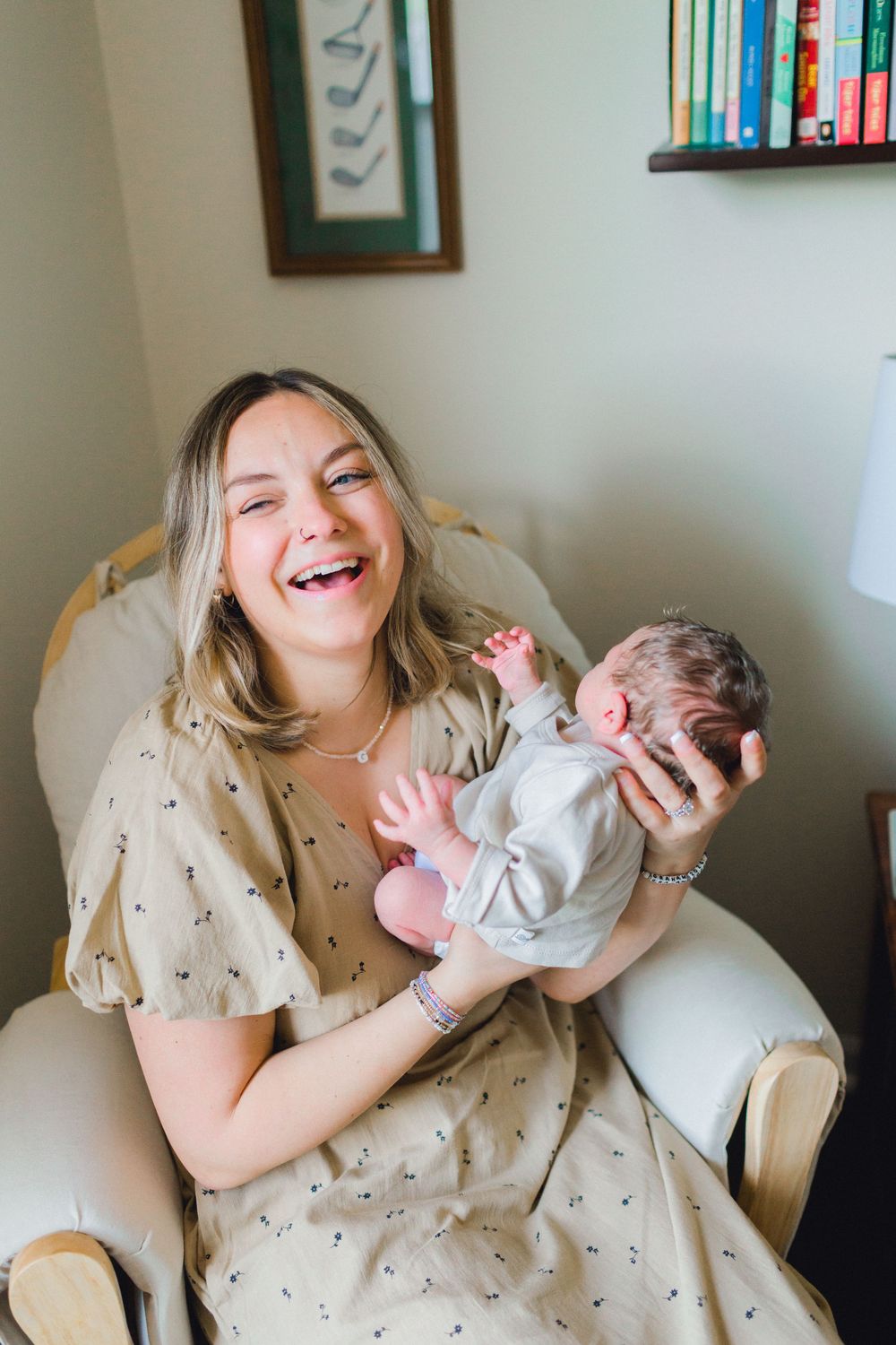 A joyful new mother laughs while holding her newborn baby in a cozy nursery chair.