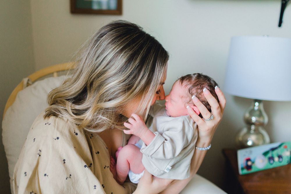 Mother tenderly kisses her newborn baby while sitting in a nursery rocking chair.