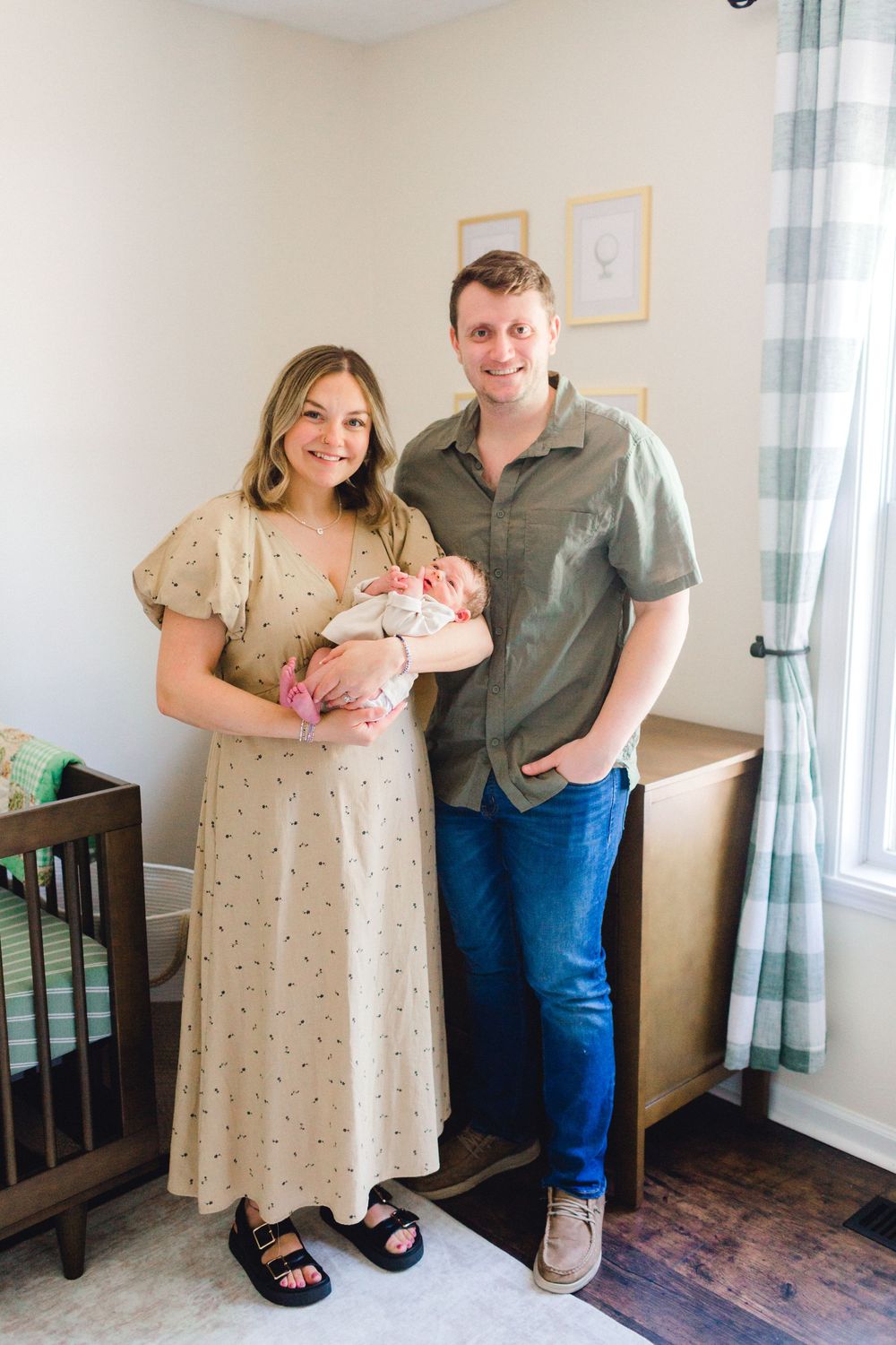 New parents smiling and holding their newborn baby in a decorated nursery with a wooden crib.
