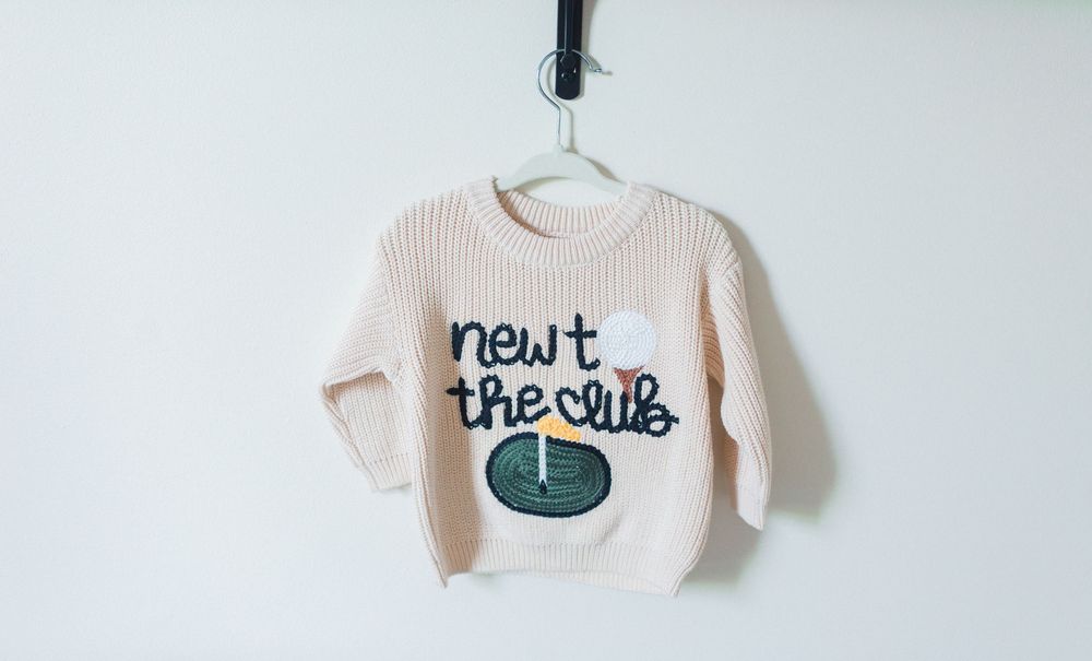 Pink knit baby sweater on hanger with 'newt the club' text and golf design applique.