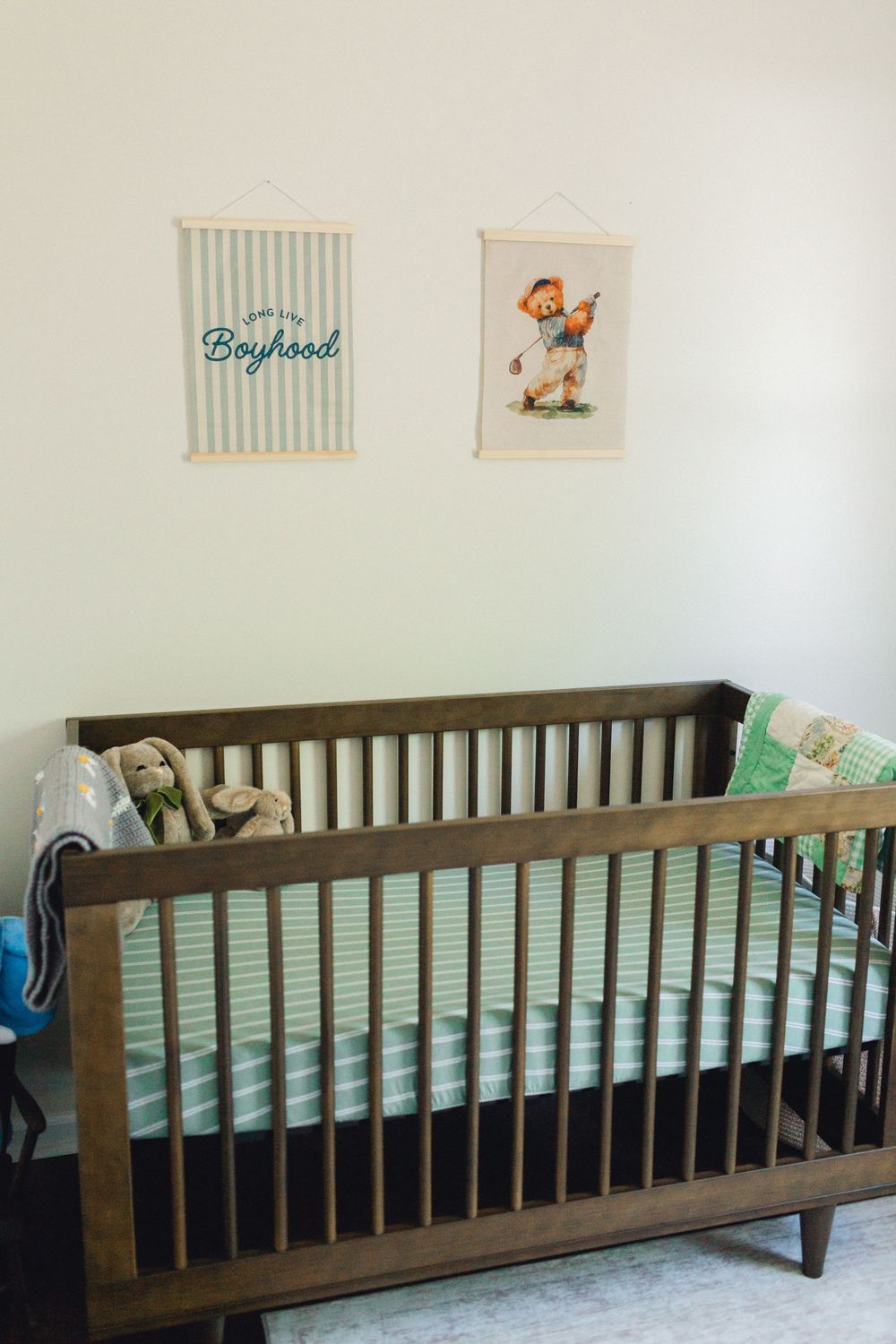 Dark wood baby crib with green striped bedding, stuffed animals, and boyhood wall art in a nursery.