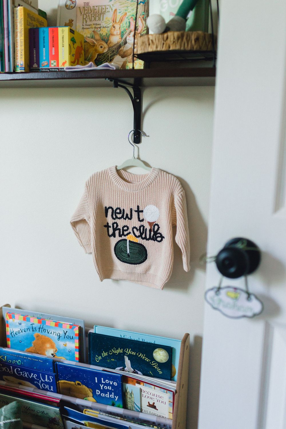 Cute pink knit baby sweater reading newt the club hanging in a decorated nursery.