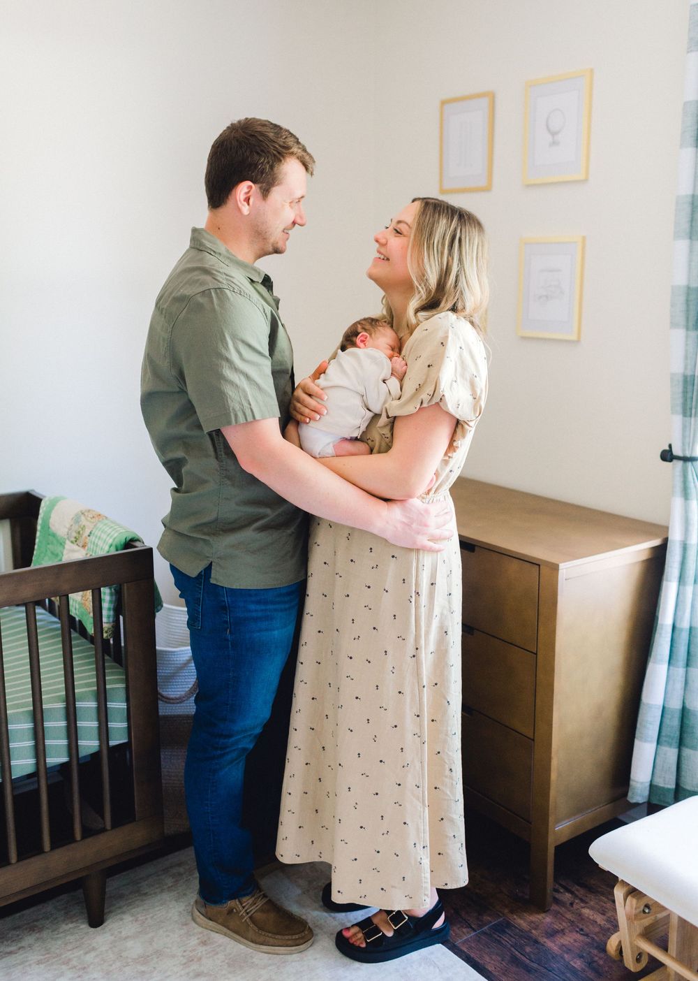 New parents smiling at each other while holding their newborn baby in a warmly decorated nursery room.