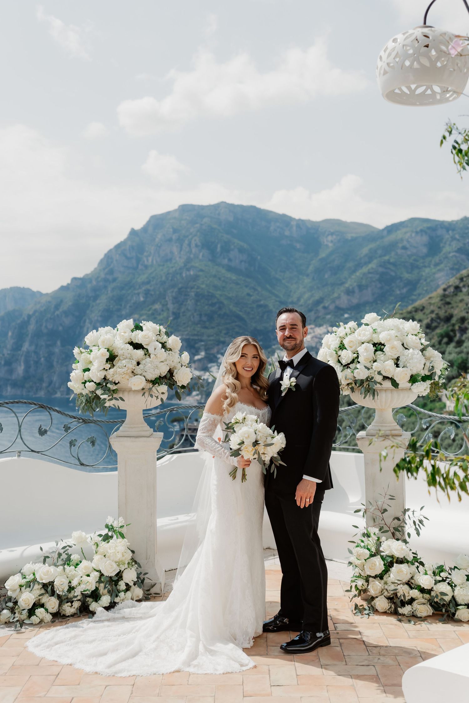Bride and groom pose on a scenic Amalfi Coast terrace with white floral arrangements and mountain views.