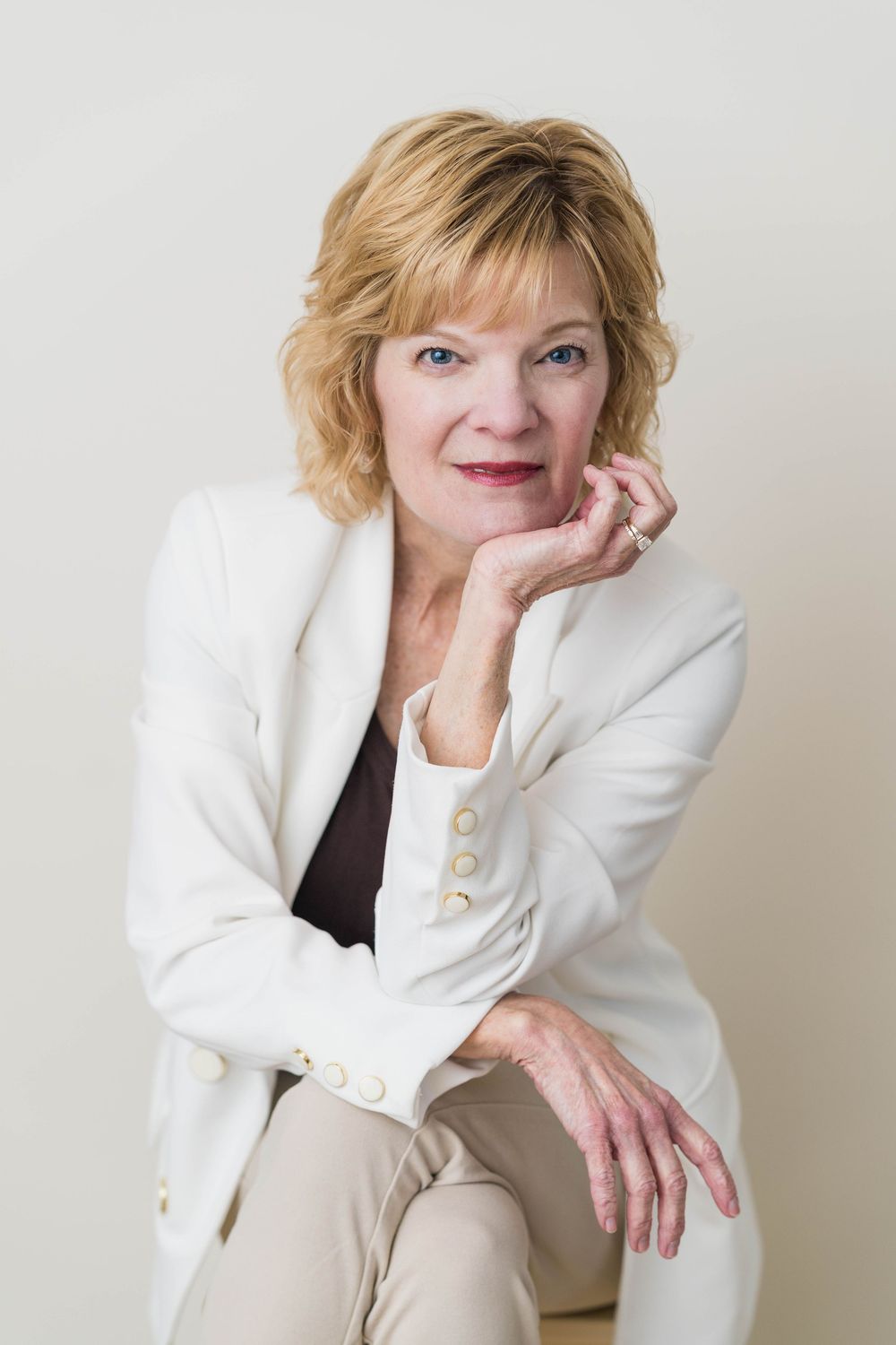 Mature blonde woman in white blazer posing confidently with hand on chin against neutral background.