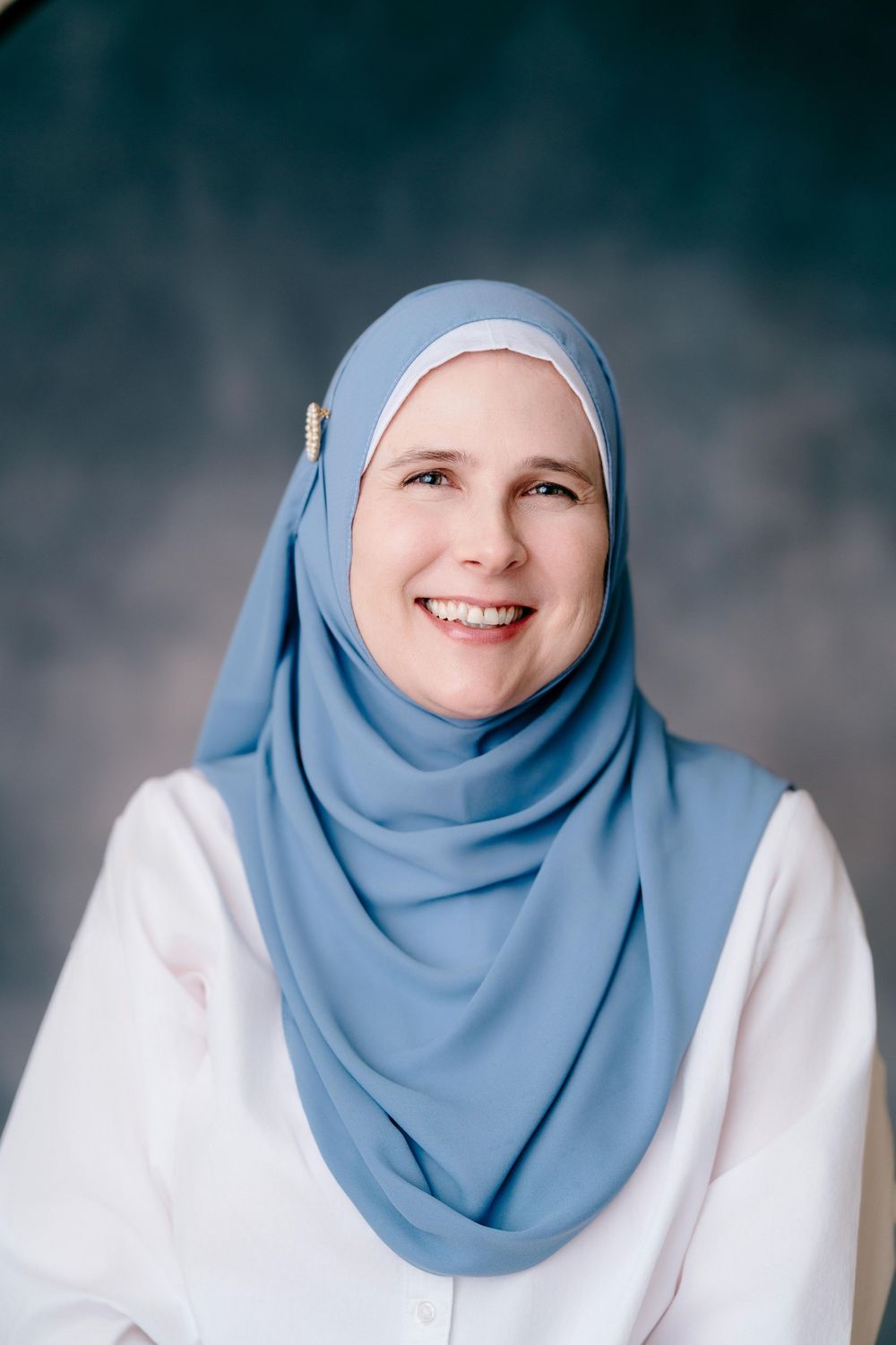 Smiling woman wearing a light blue hijab and white top poses for a portrait against a dark gray background.