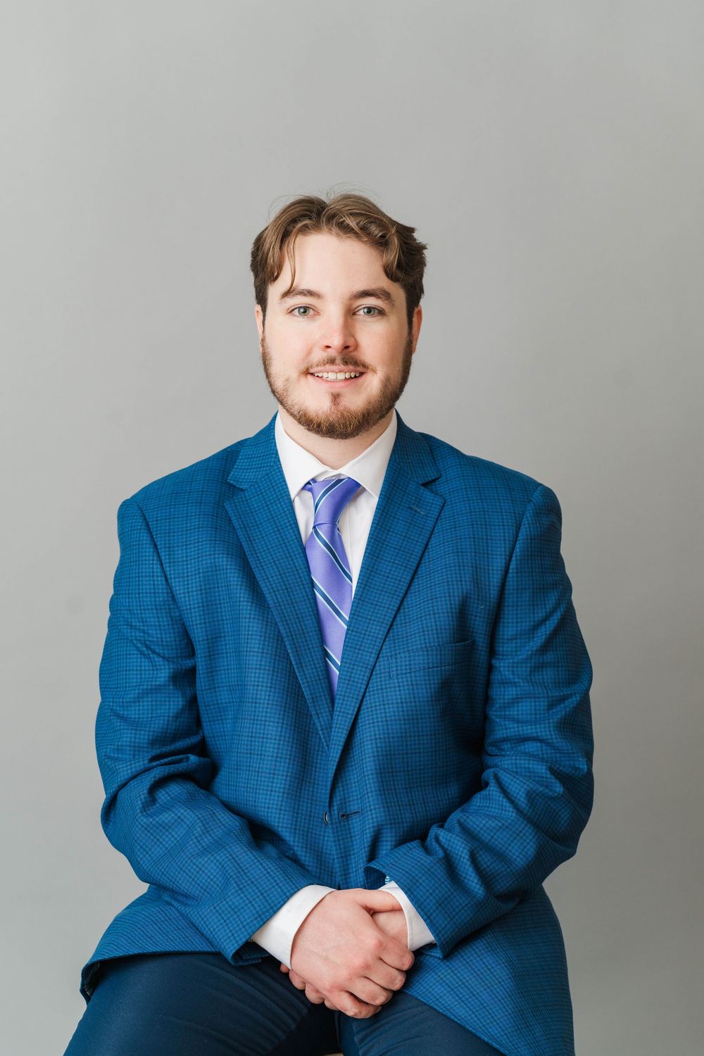 Young professional man in blue suit and purple tie smiling, seated against gray background.