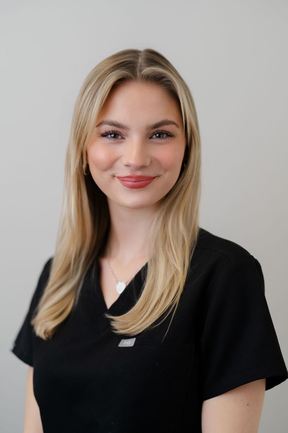 Young blonde woman in black scrubs smiling professionally against a light gray background.