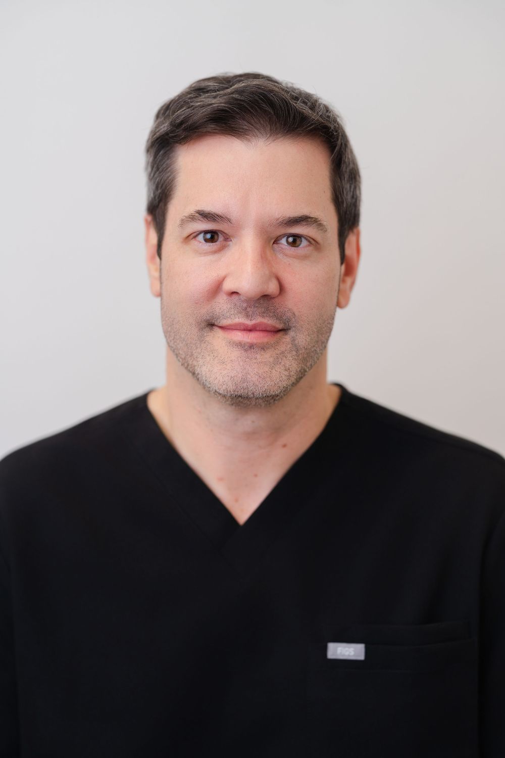 A male healthcare professional in black scrubs poses for a professional headshot against a light gray background.