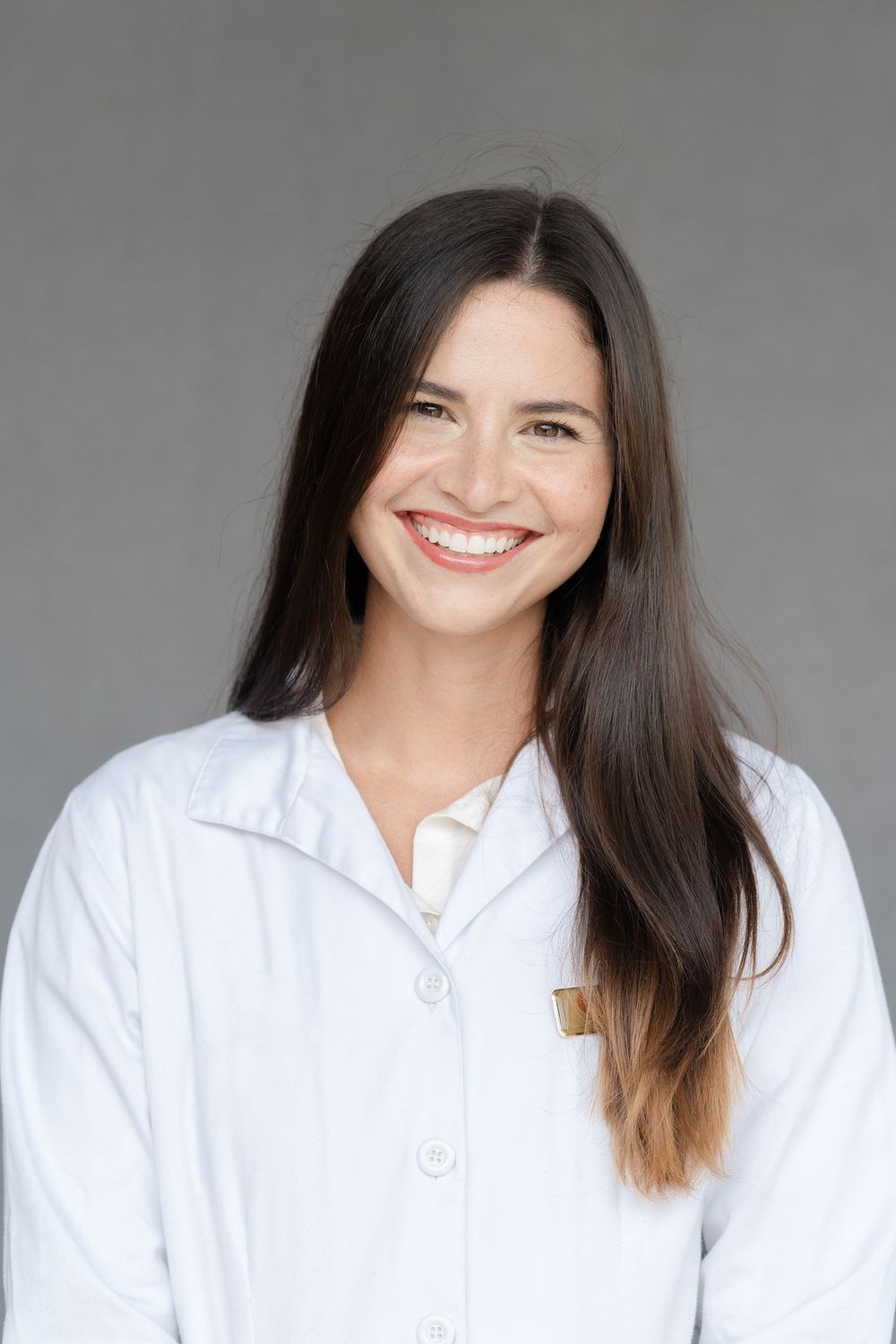 Smiling young woman with long dark hair wearing a white lab coat and name badge against a gray background.