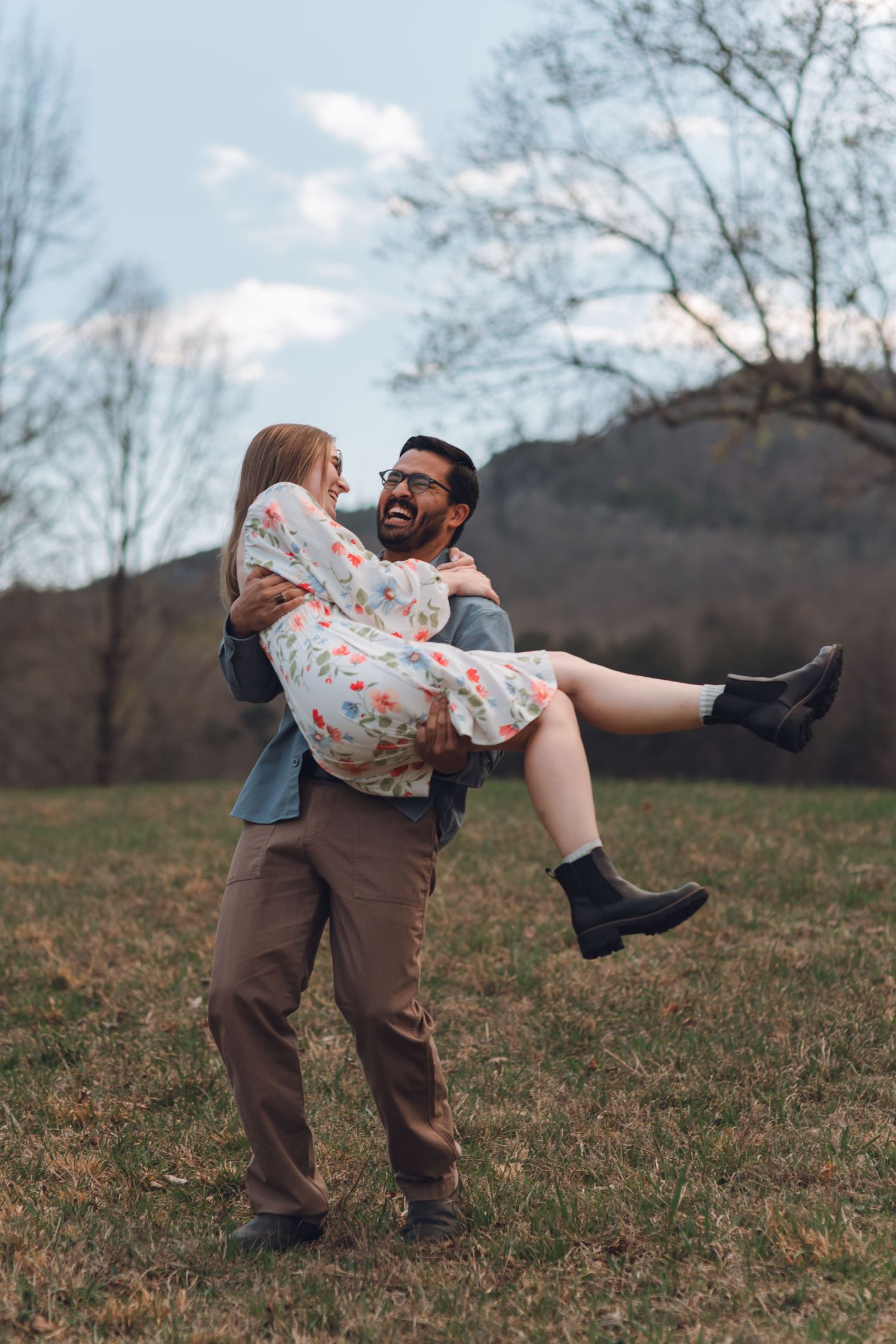 A joyful man carries a laughing woman in a floral dress outdoors in a grassy field with trees and mountains.