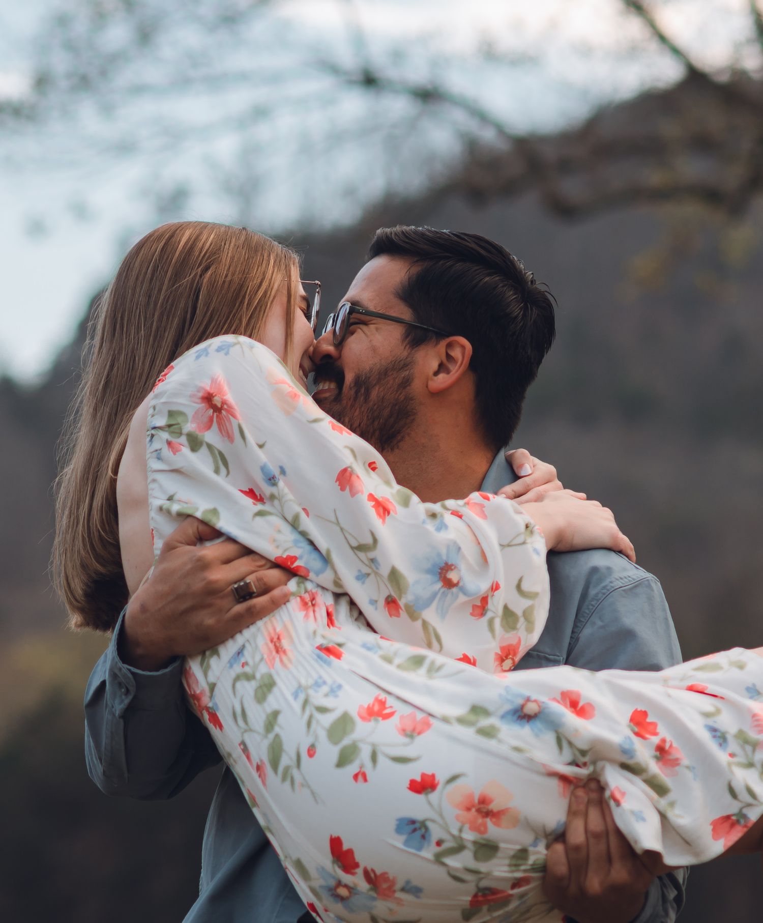 A man in a gray jacket lifts and kisses a woman in a floral dress outdoors with trees in the background.