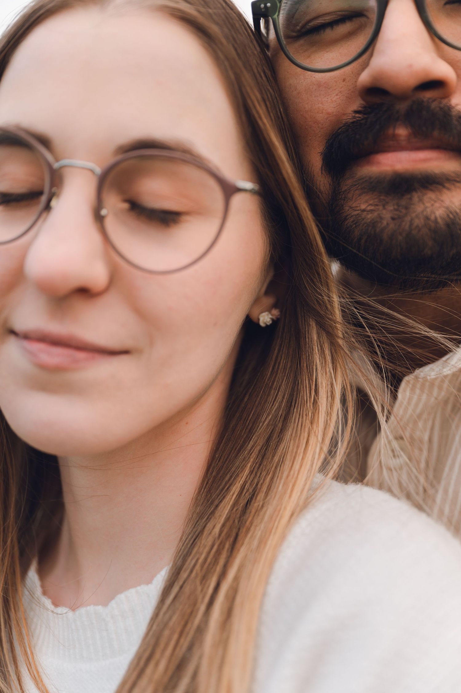 Close-up portrait of a couple with glasses, woman with long blonde hair in white sweater, man with beard leaning close.