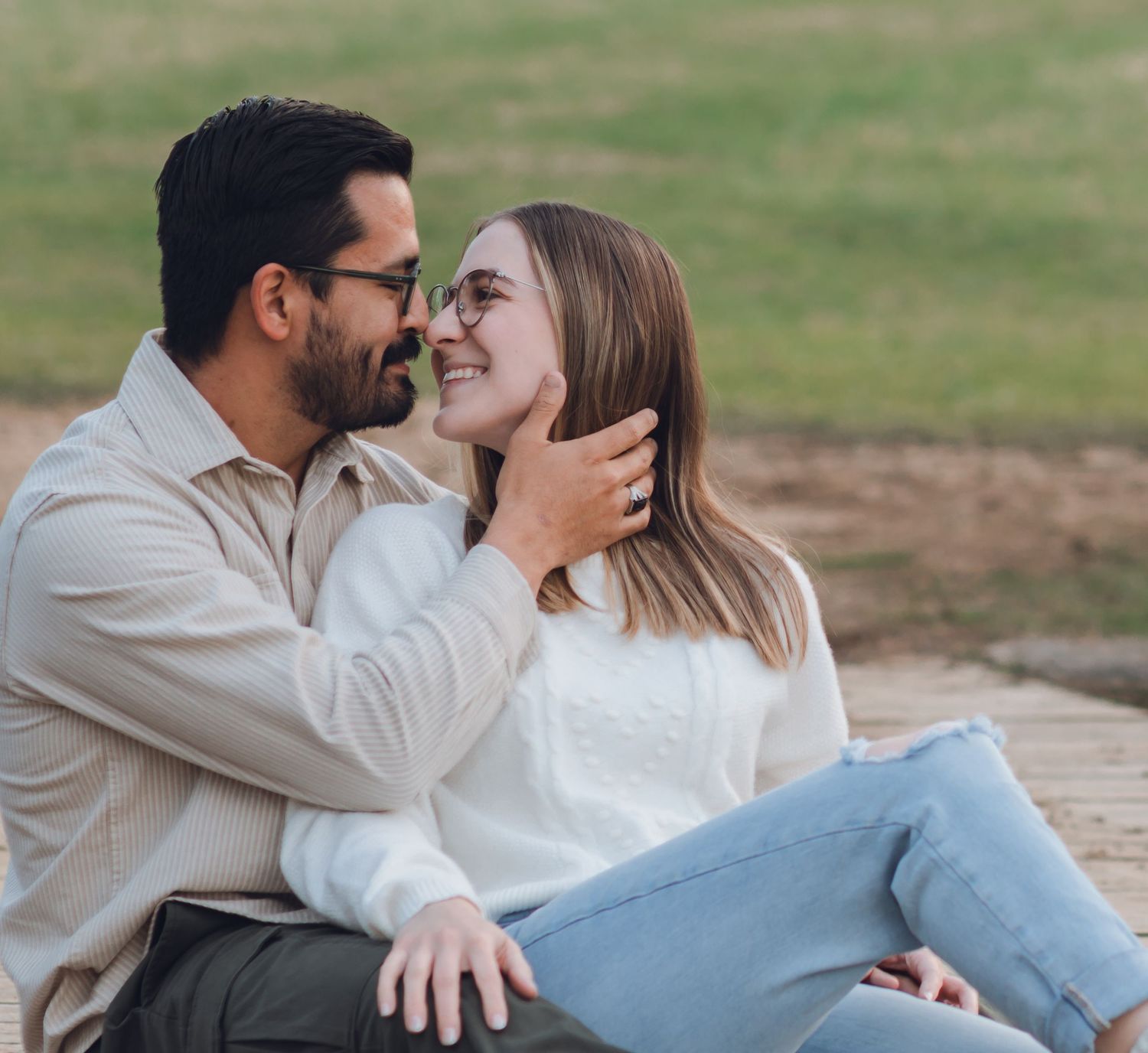 A couple sitting outdoors, sharing a romantic moment, smiling and gazing at each other while the woman touches the man's face.