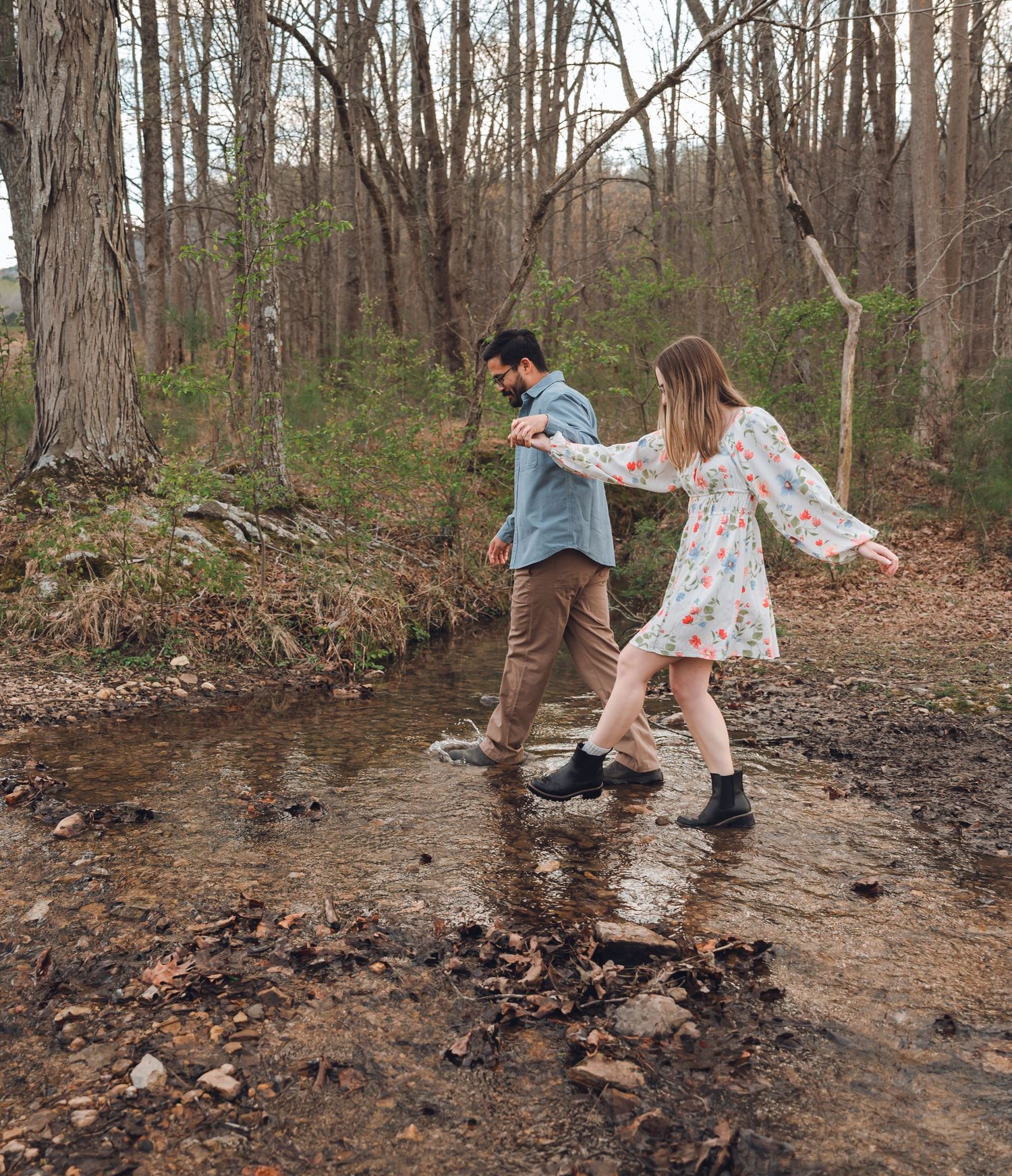 Couple crossing a rocky stream in a forest, woman in floral dress and man in blue shirt holding hands.