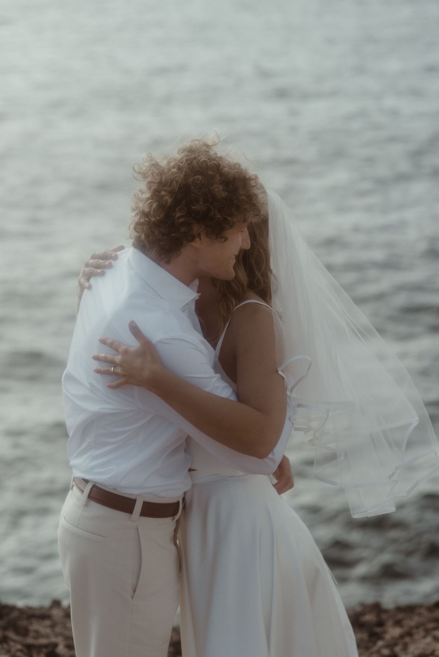 Bride in white gown and veil walks toward groom in beige suit on scenic coastal garden path overlooking the sea.