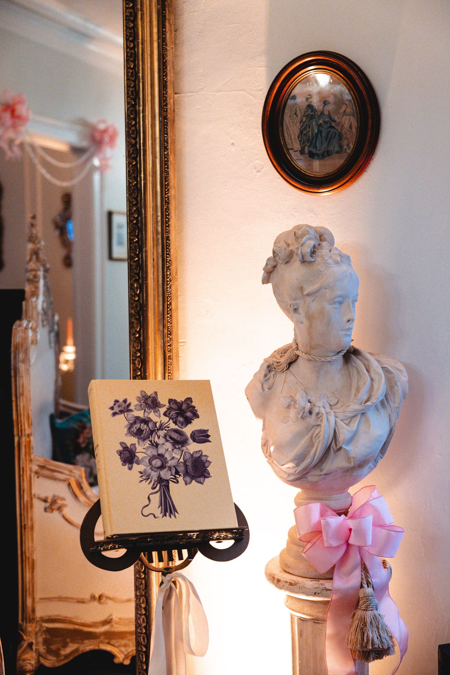 Elegant antique decor featuring a white marble bust with pink bow, ornate gold mirror, and vintage floral artwork on display stand.