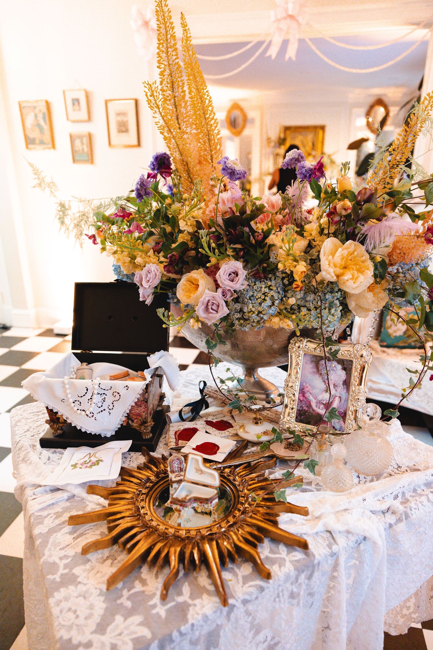 Elegant wedding reception table with lush floral arrangement, gold sunburst mirror, framed photos, and lace tablecloth.