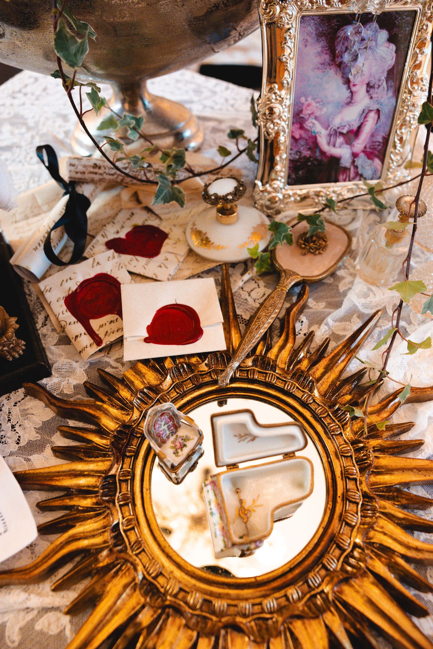 Ornate gold sunburst mirror on vintage lace table with antique portrait, wax seal letters, and decorative trinkets.