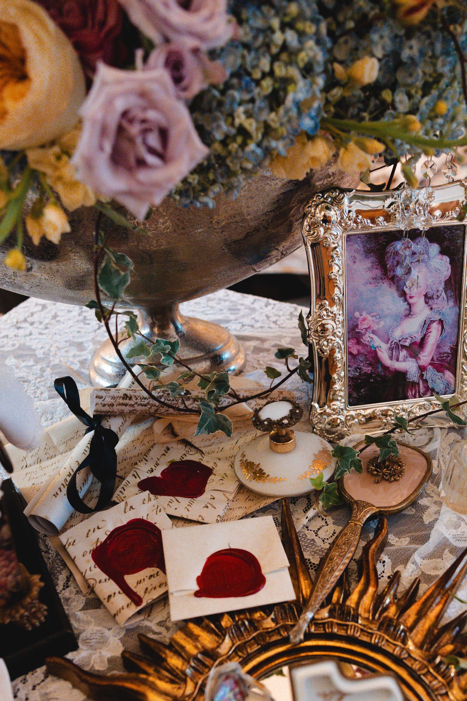 Vintage tableau with roses, wax seal letters, ornate gold frame portrait, lace, and antique mirror on decorative table.