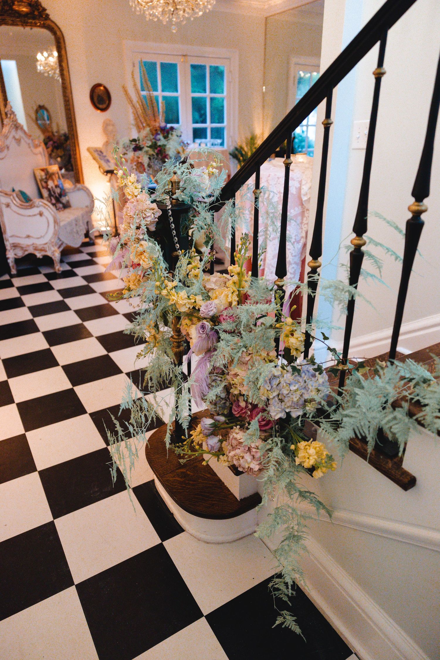 Elegant foyer with black and white checkered floors, ornate staircase adorned with lush floral arrangement and gilded mirror.