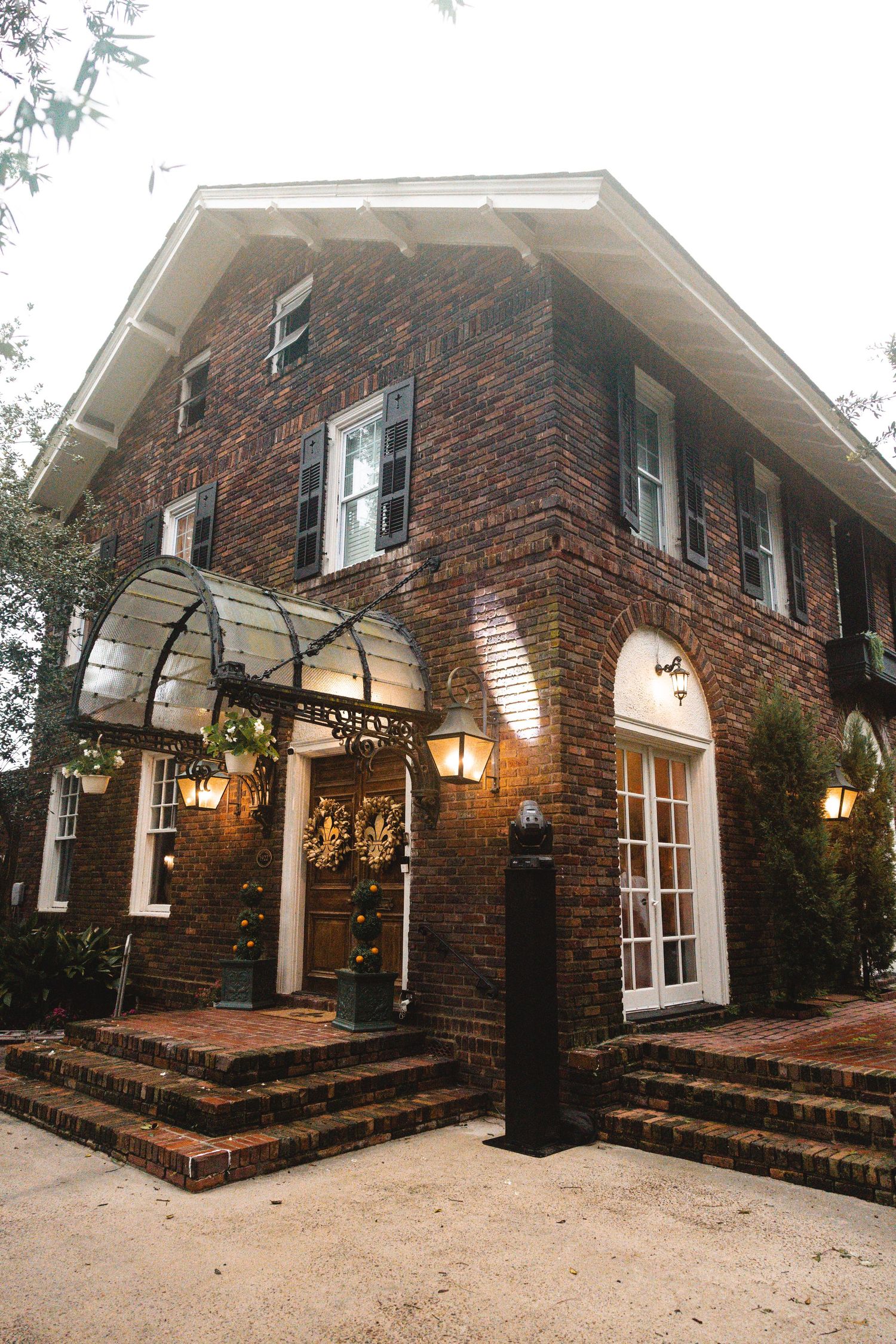 Charming two-story brick building with arched entrance canopy, glowing lanterns, and French doors surrounded by lush greenery.