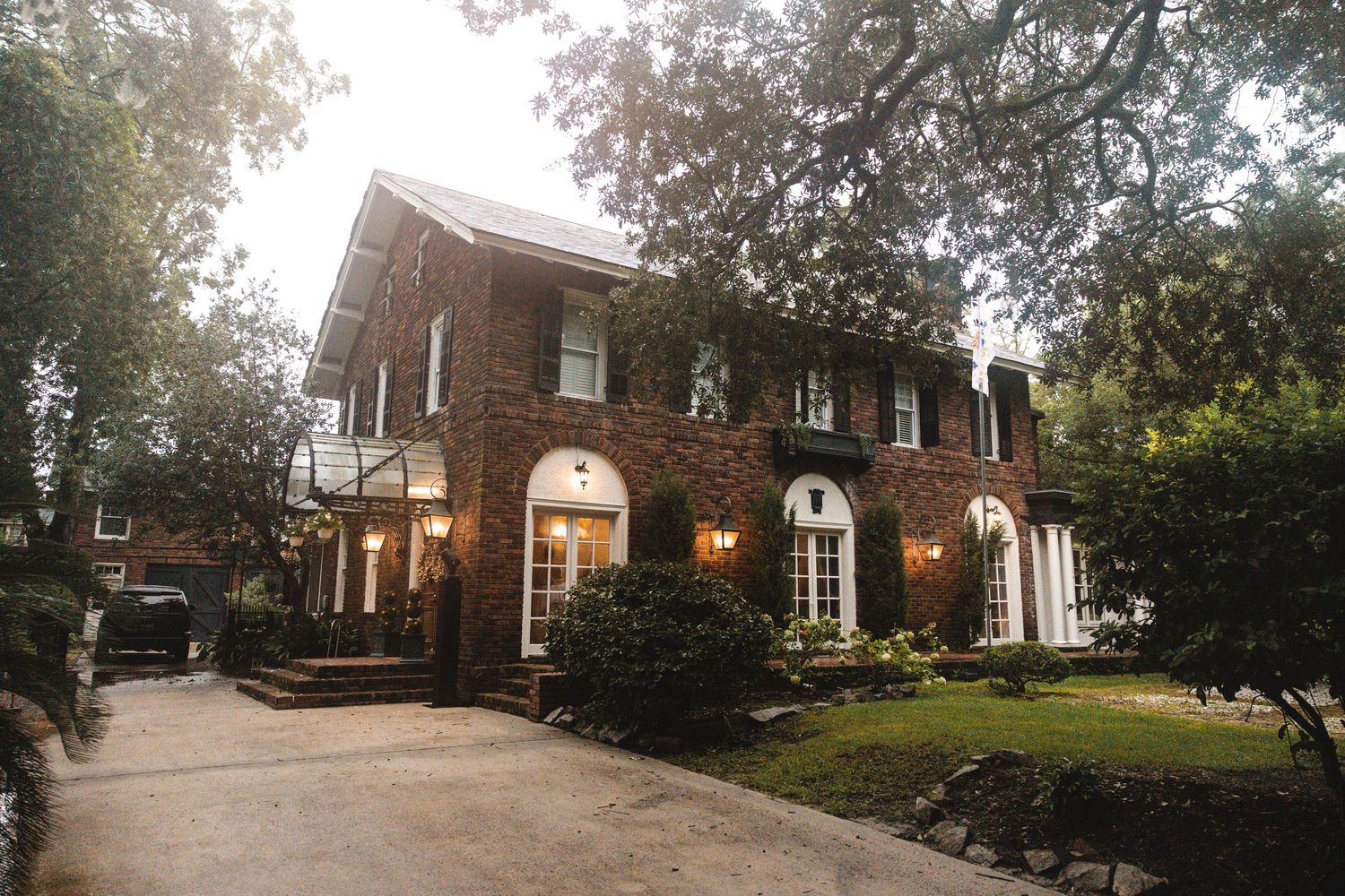 A charming two-story brick colonial home surrounded by lush trees with arched windows and a paved driveway.