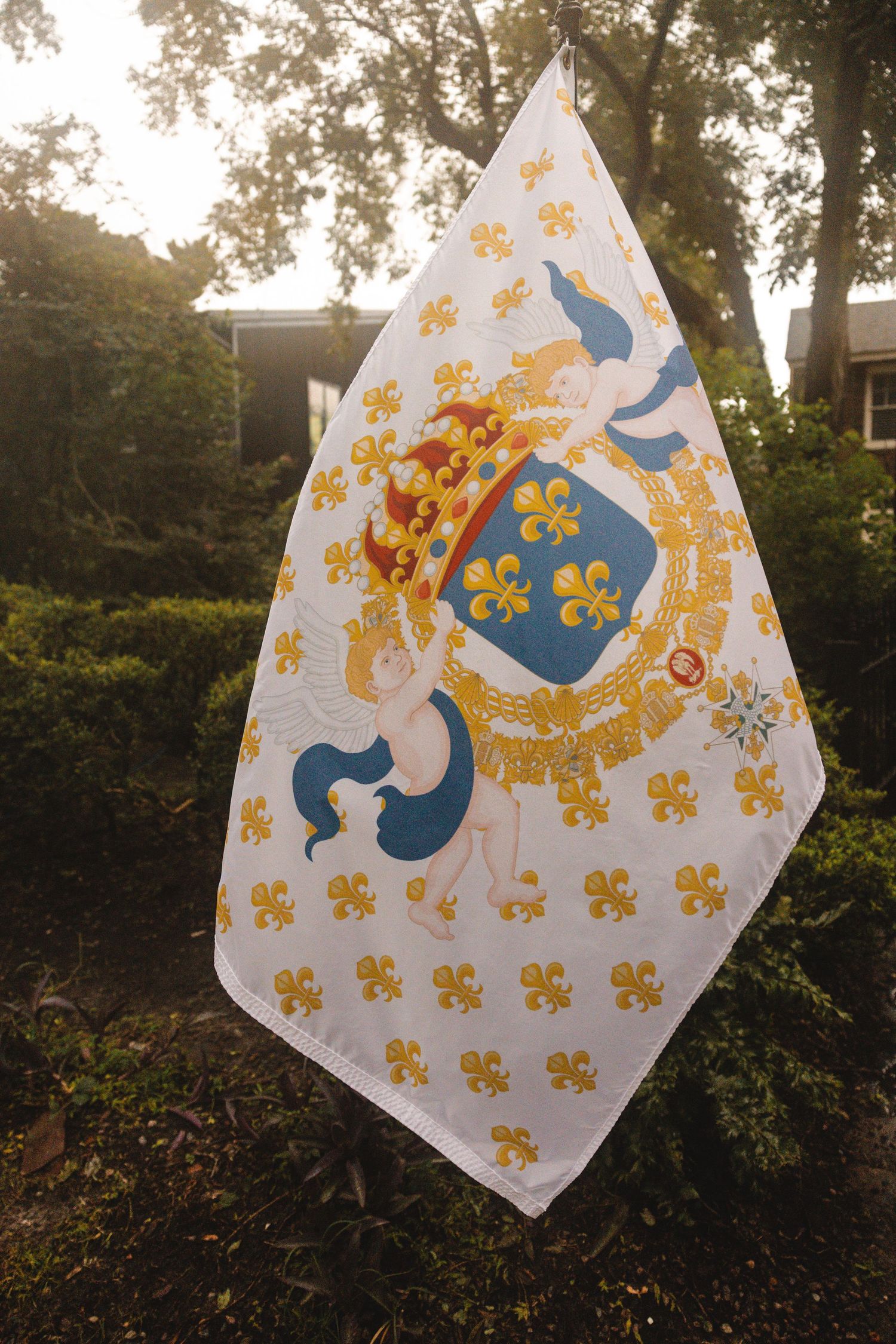 White heraldic flag with gold fleur-de-lis pattern and blue coat of arms shield displayed outdoors among green trees.