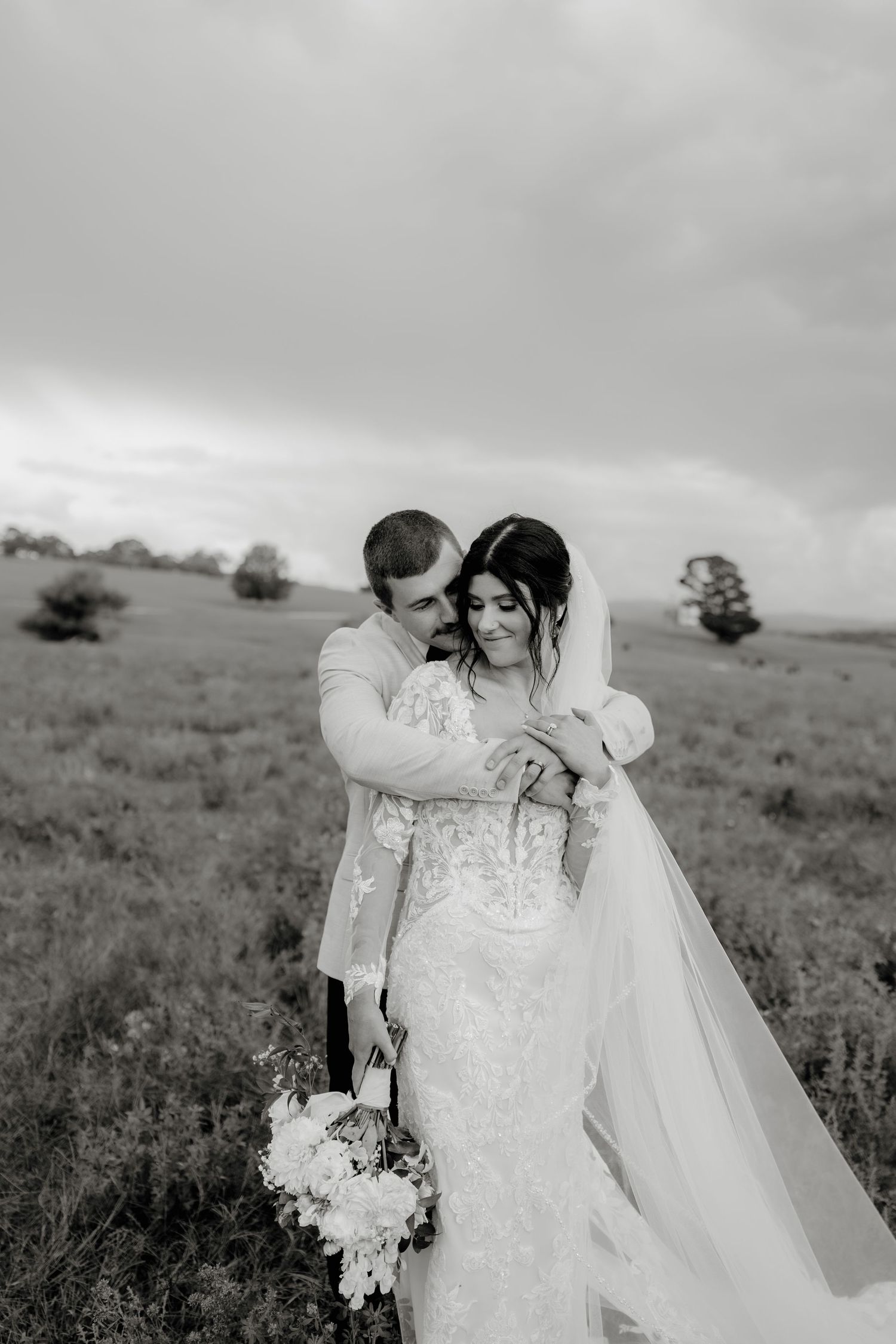 Black and white photo of a bride and groom embracing in a field, bride holding a floral bouquet wearing a lace gown.