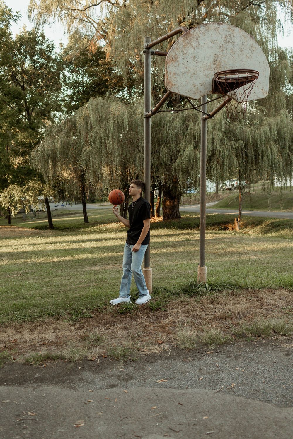 A basketball player in casual attire practices their jump shot at an outdoor court with willow trees in the background.