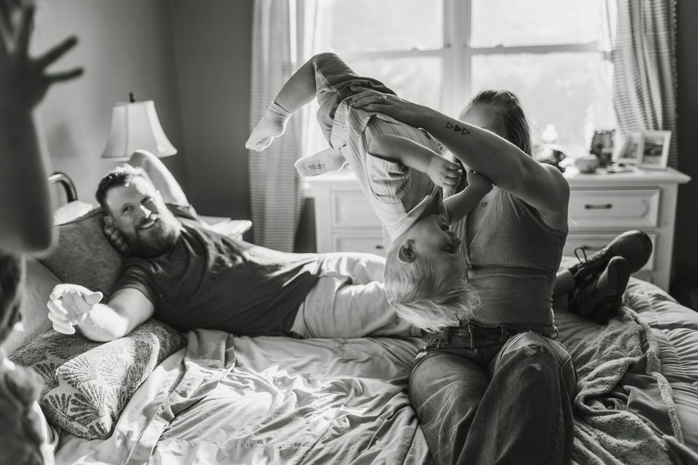 Black and white photo of a family playing on a bed, with a man lying down and another lifting a baby joyfully.