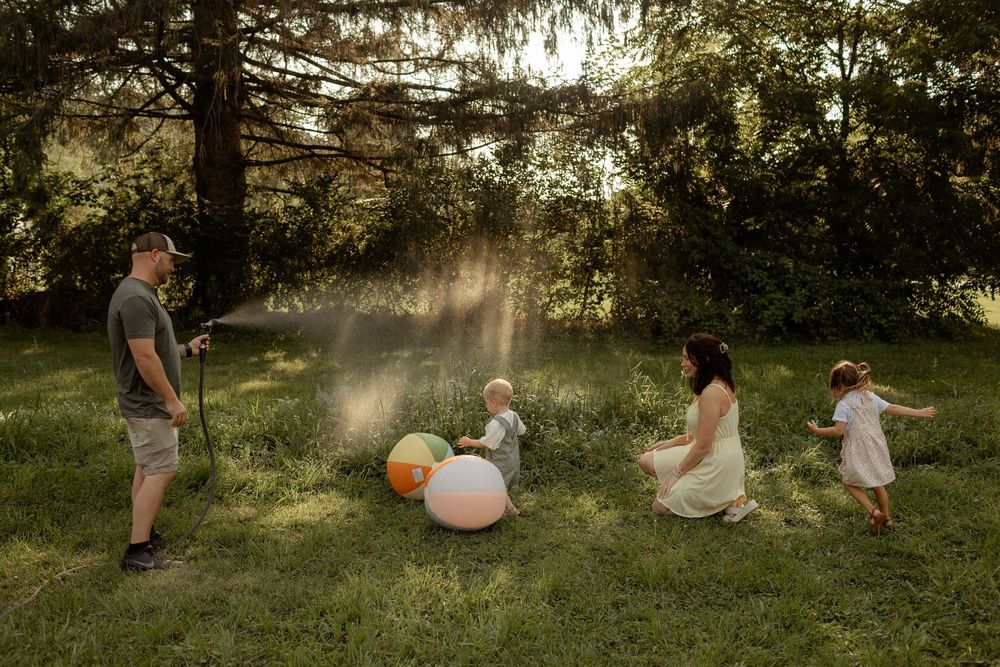 Family playing outdoors in sunlit yard, father spraying water hose while children and mother enjoy colorful balloons on grass.