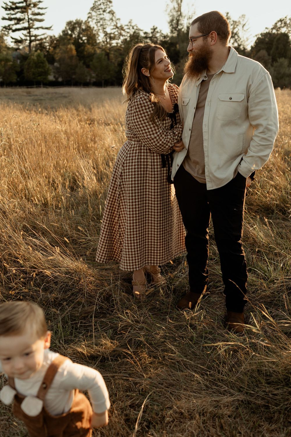 A couple shares a tender moment in a golden field at sunset while a small child plays in the foreground.