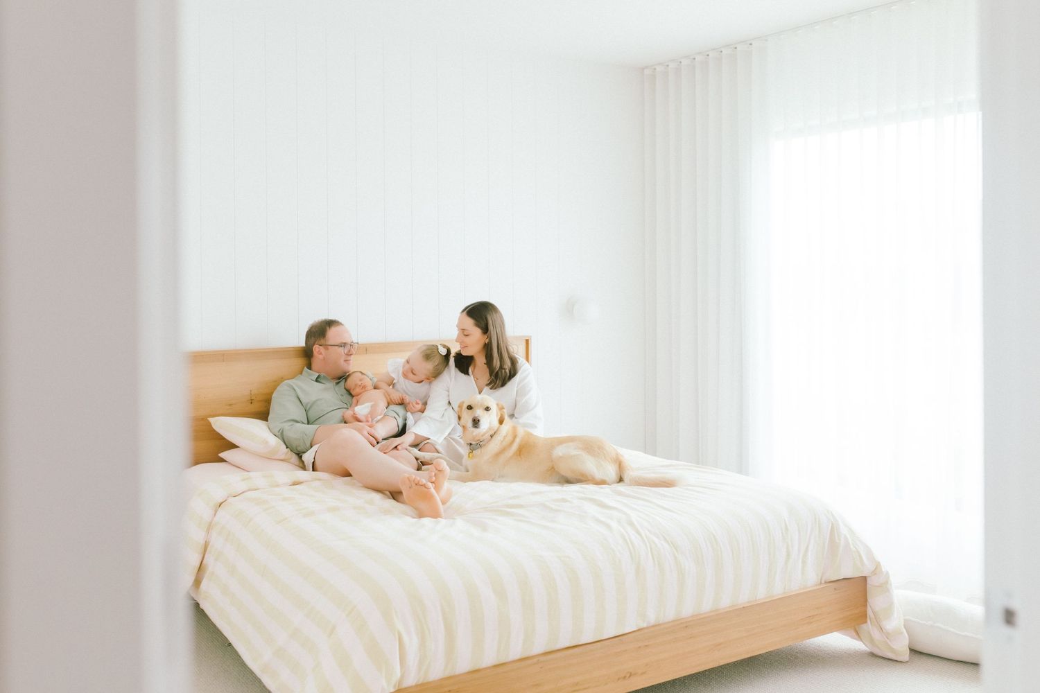 Family relaxing on a modern bed with a golden retriever dog in a bright white minimalist bedroom.