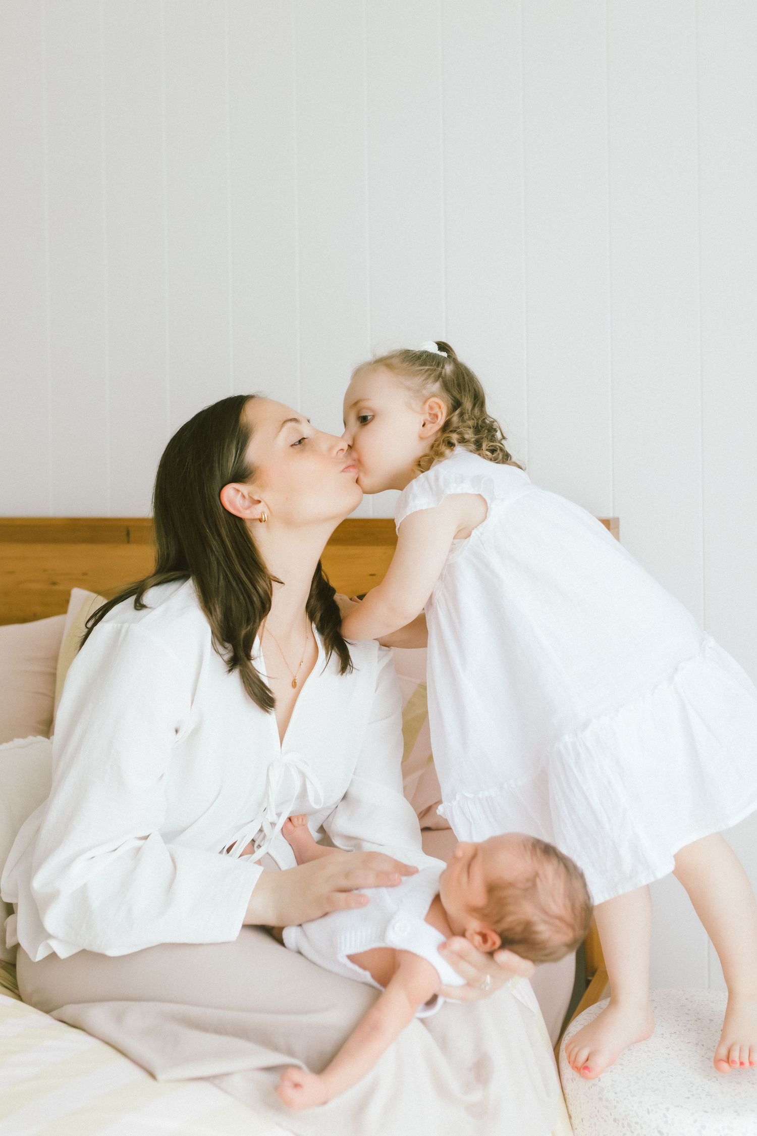 A mother in white holds her newborn while her young daughter in a white dress gives her a sweet kiss on the cheek.