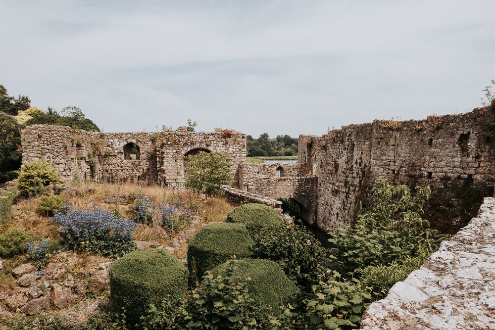 Ancient stone castle ruins with manicured topiary hedges and wildflowers under a cloudy sky.