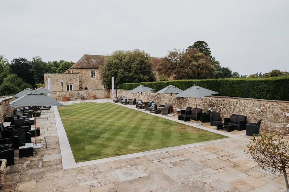 Elegant castle courtyard venue with manicured lawn, stone walls, and outdoor seating with grey parasols.