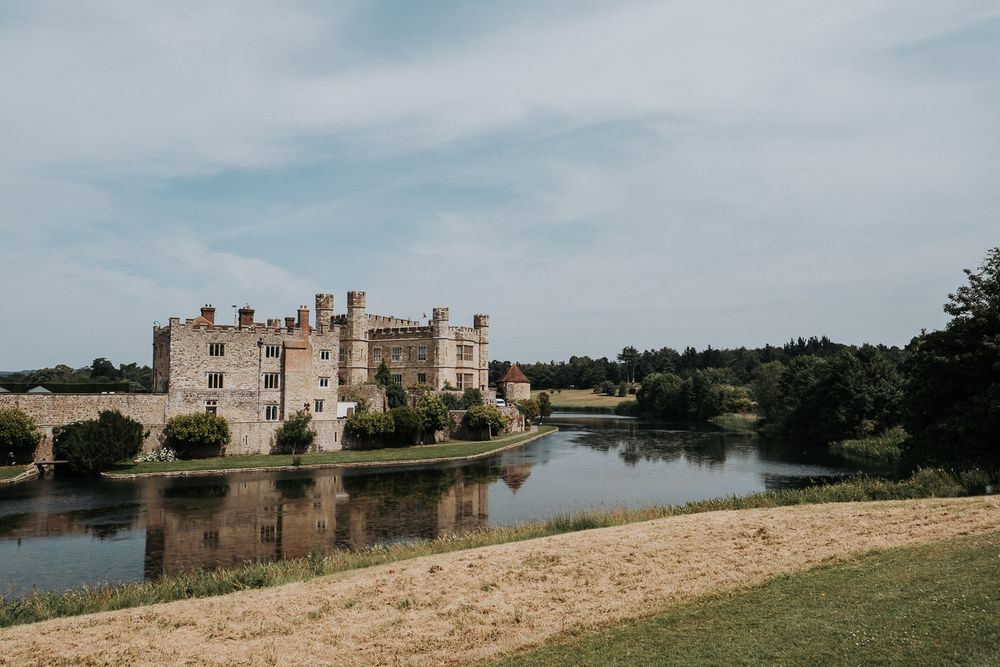 Leeds Castle reflected in its tranquil moat, surrounded by lush greenery under a partly cloudy sky in Kent, England.