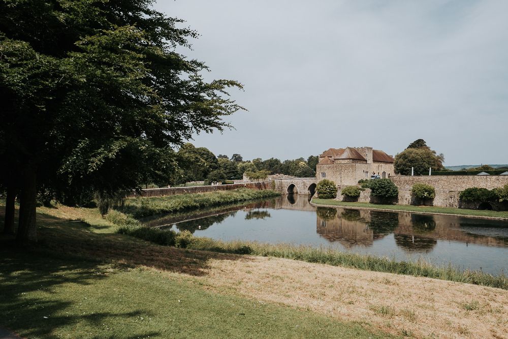 Historic castle ruins reflected in a serene moat surrounded by lush green trees and manicured lawns on a cloudy day.