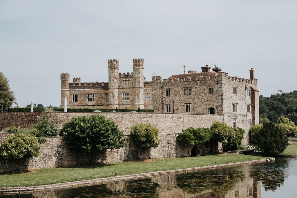 Historic Leeds Castle with medieval stone walls and battlements reflected in a serene moat, surrounded by lush greenery.
