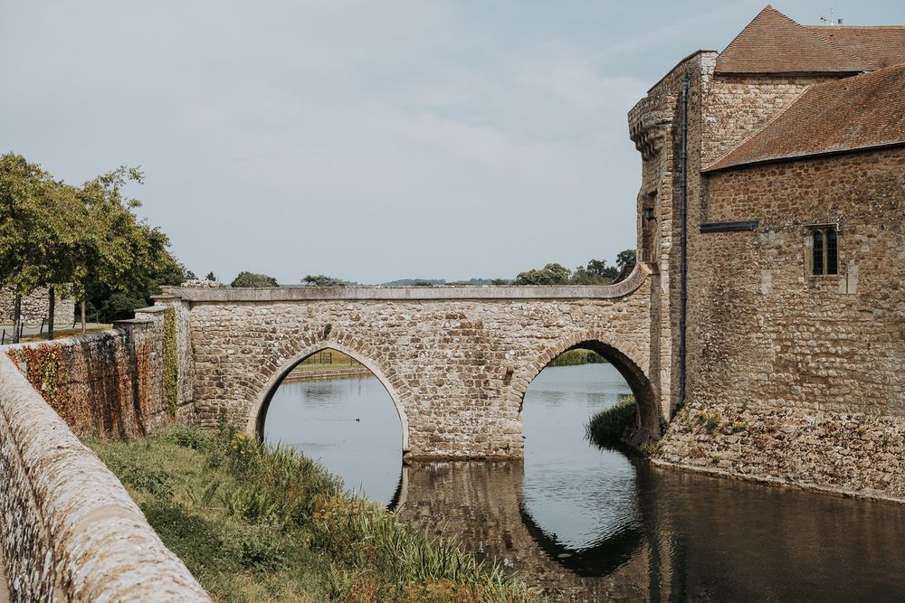 Historic stone bridge with arched openings spanning the moat at Leeds Castle, Kent, England, under a clear sky.