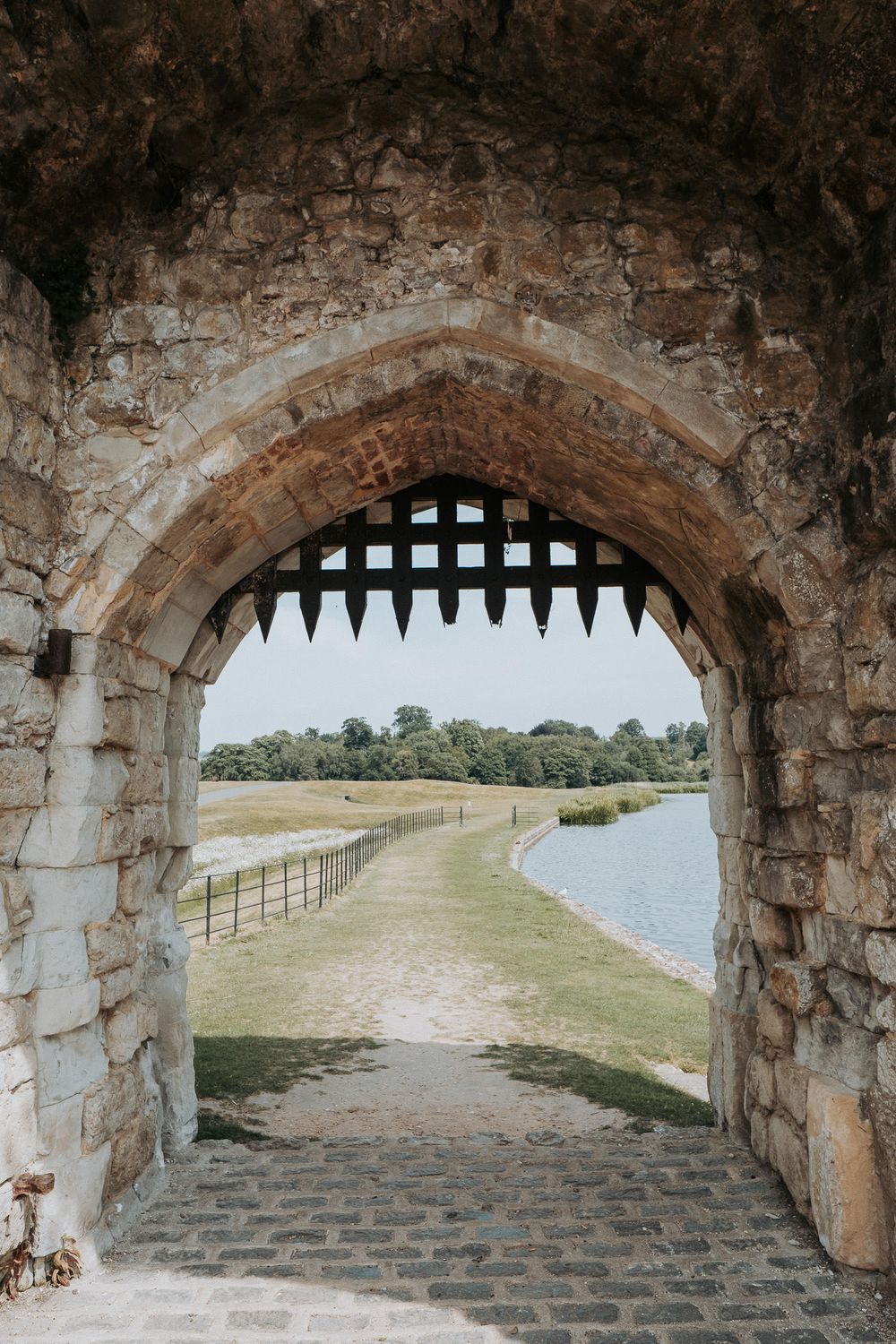 Medieval castle archway with portcullis framing a scenic view of a river path and lush green trees beyond.