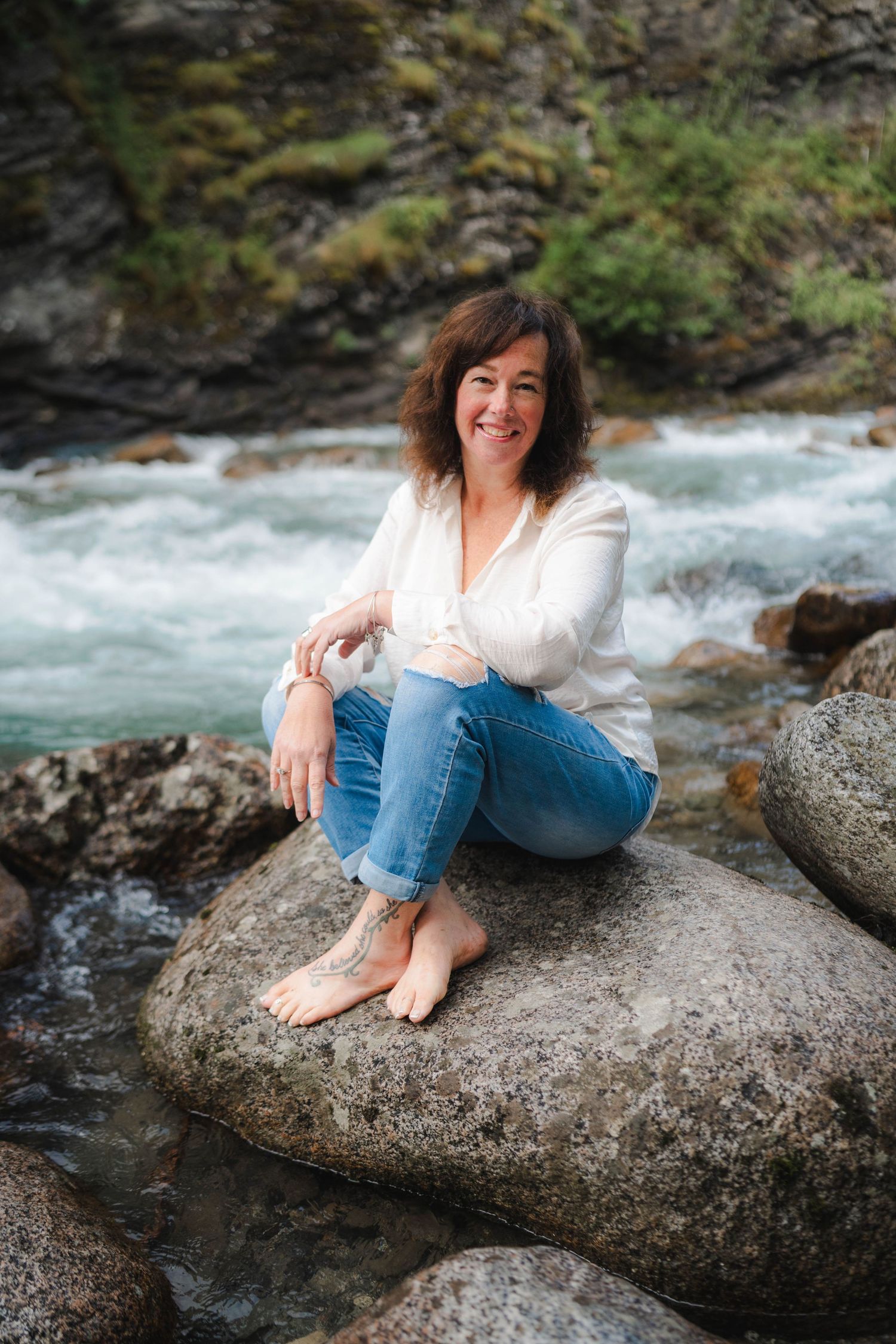 A smiling woman sits barefoot on river rocks in jeans and white top, with a rushing mountain stream behind her.