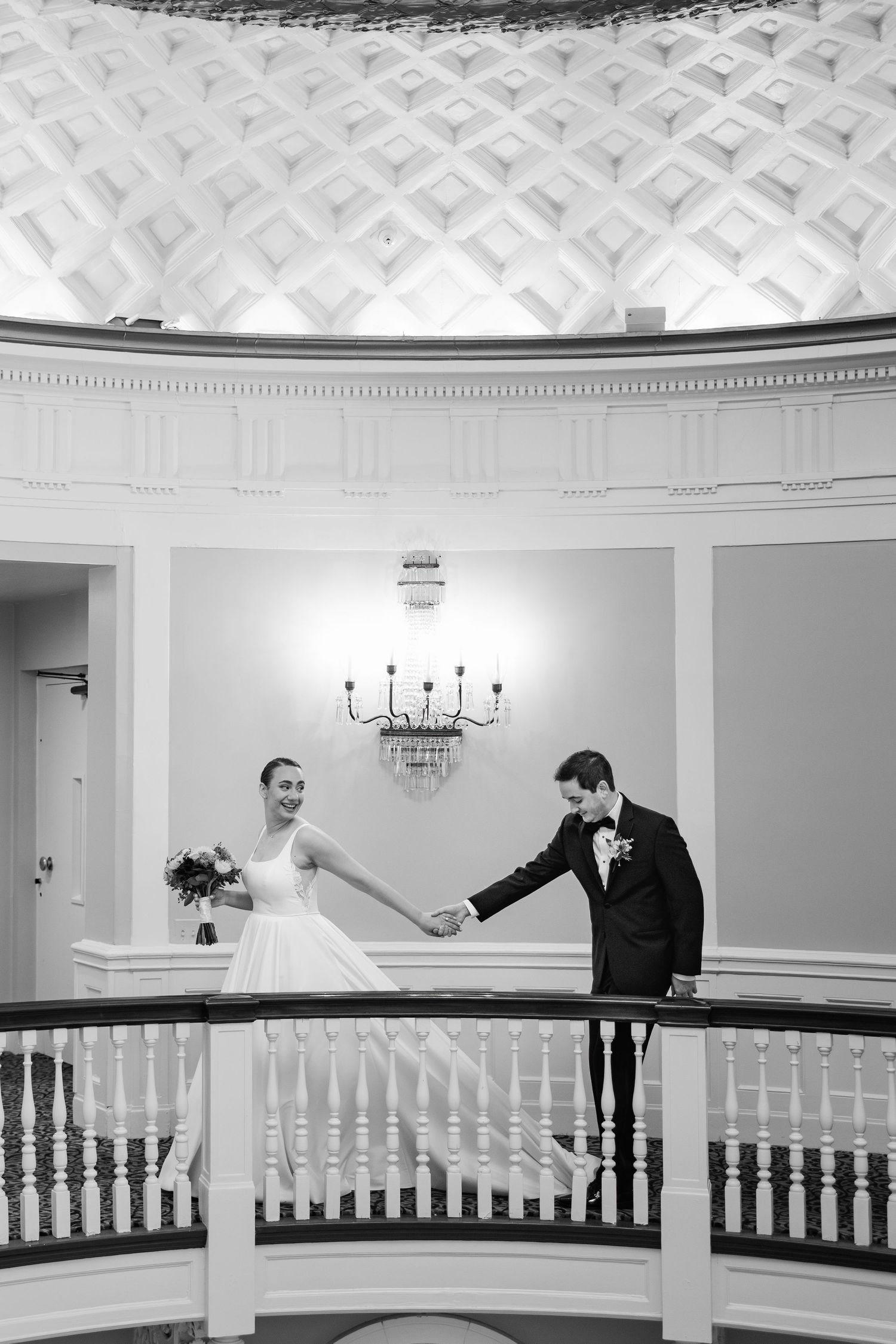 Black and white photo of bride and groom holding hands on elegant balcony with ornate ceiling.