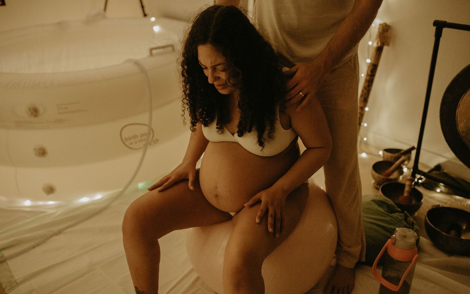 Pregnant woman sitting on birthing ball smiling, receiving back support during labor preparation in cozy room.