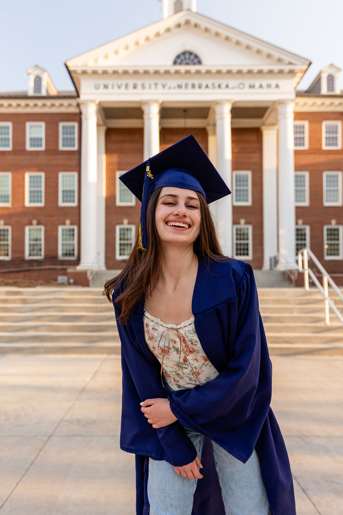 Smiling young woman in navy blue graduation cap and gown standing in front of a University of Nebraska Omaha building.